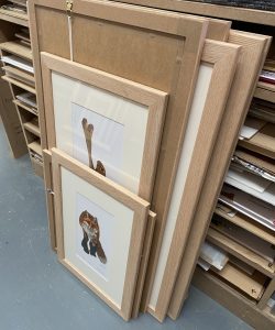 A stack of wooden framed images leaning against some storage on a grey painted workshop floor